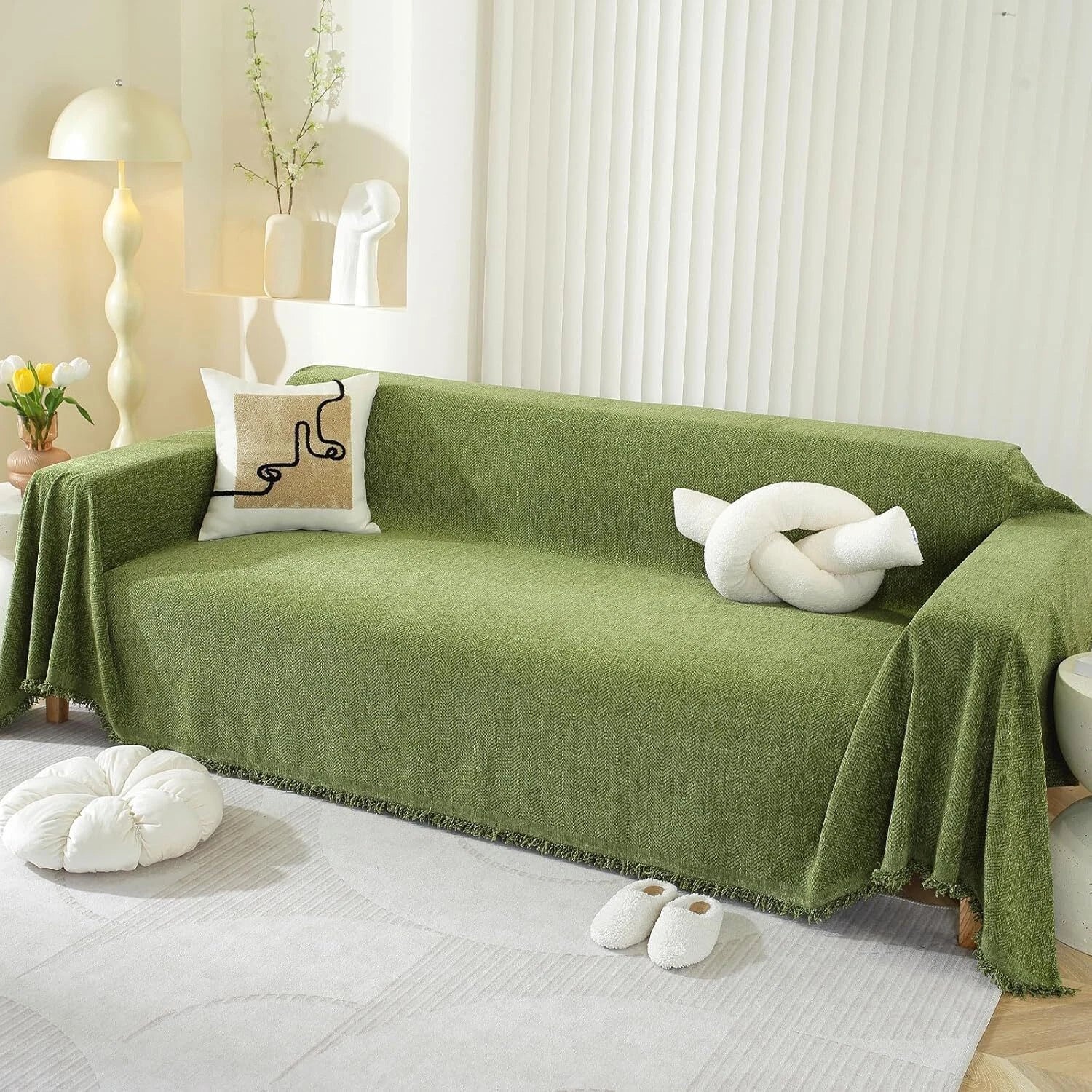 An olive green textured sofa slipcover draped over a three-seat sofa in a bright, modern living room setting, styled with decorative pillows and a white knotted throw pillow.